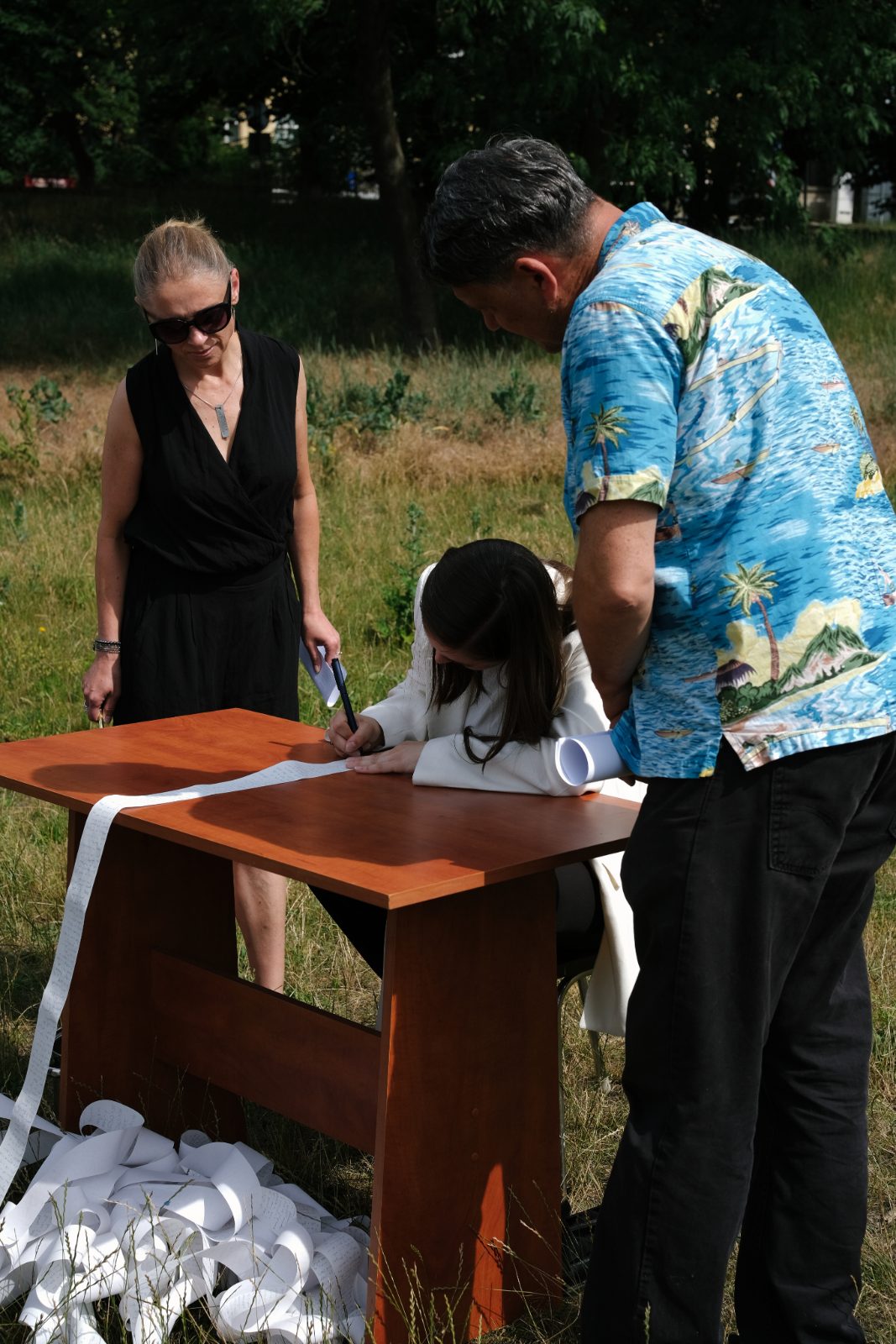 A woman, a man in a Hawaiian shirt and a girl signing a scroll at a table outdoors
