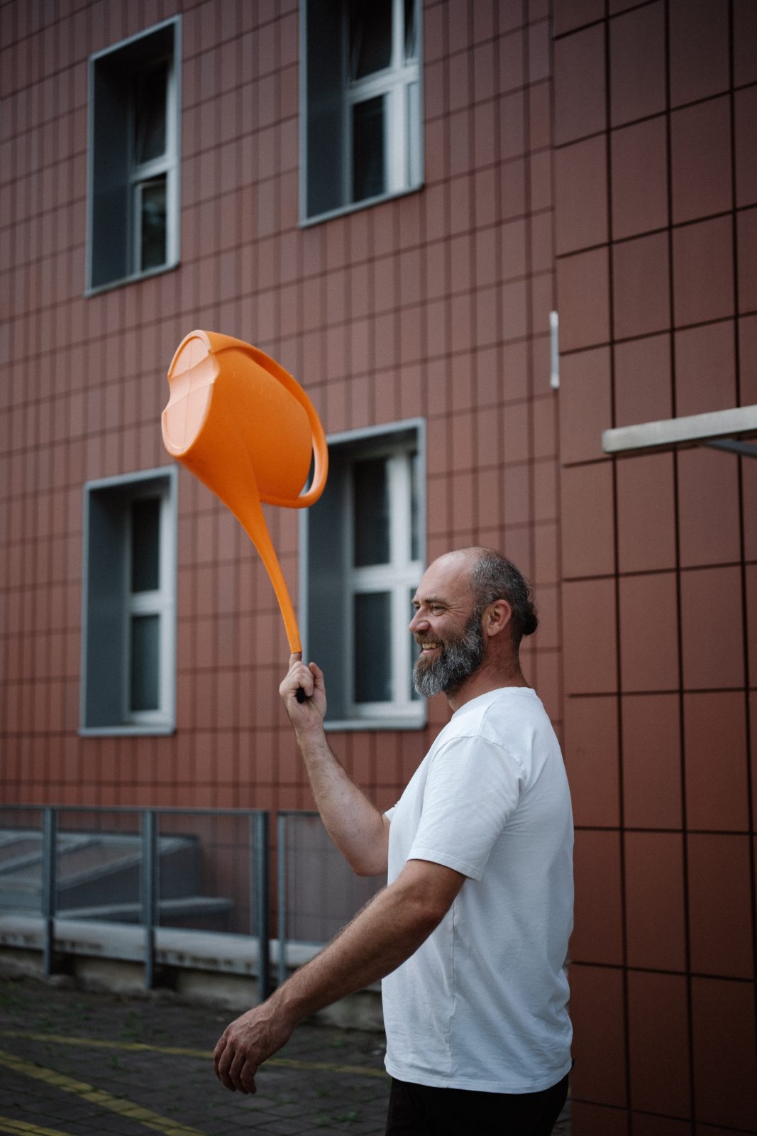 A laughing man pouring from an orange watering can in front of a brick building