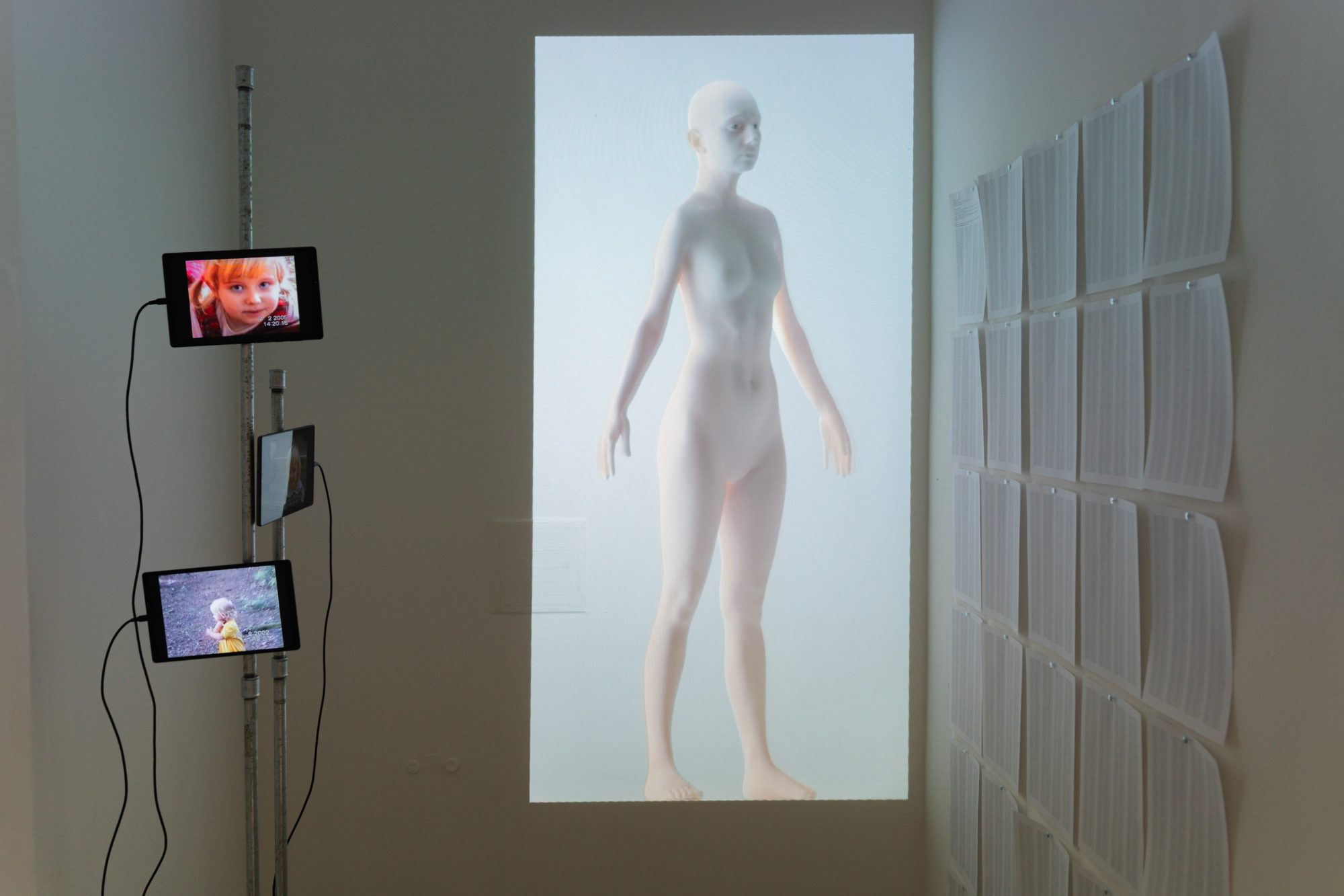 Installation view: tablets with childhood VHS footage, a projected digital body and a wall of printed sheets
