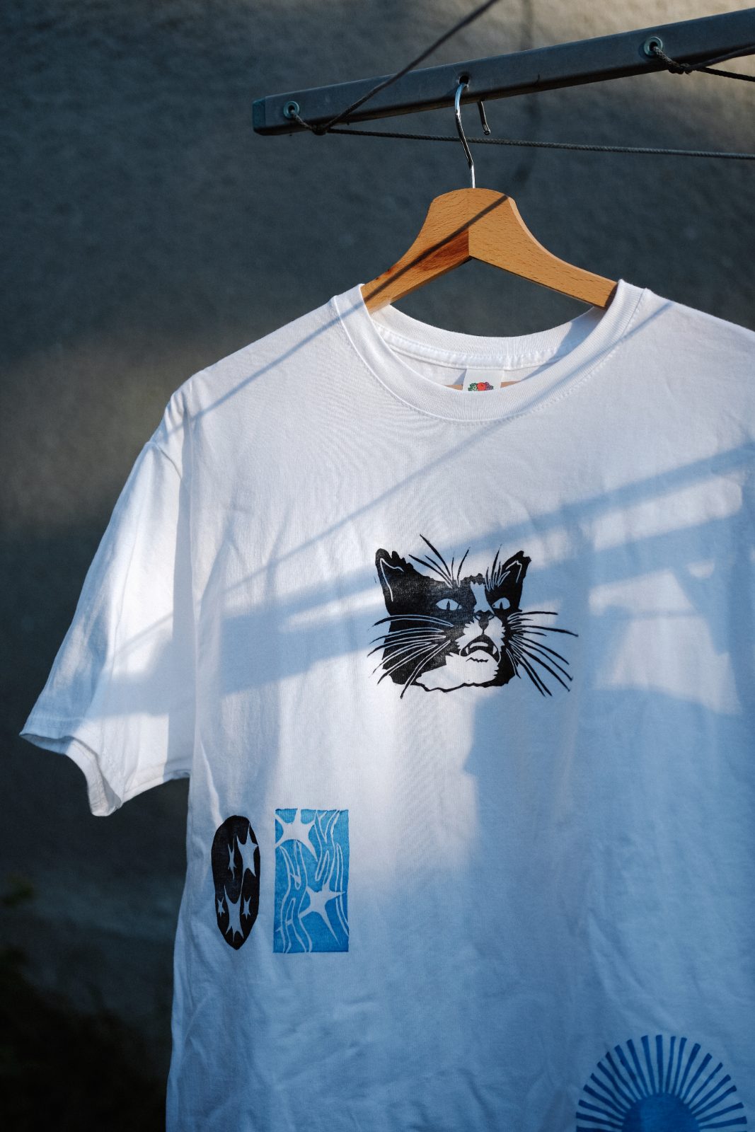 White T-shirt with a linocut cat portrait and small star prints, on a hanger